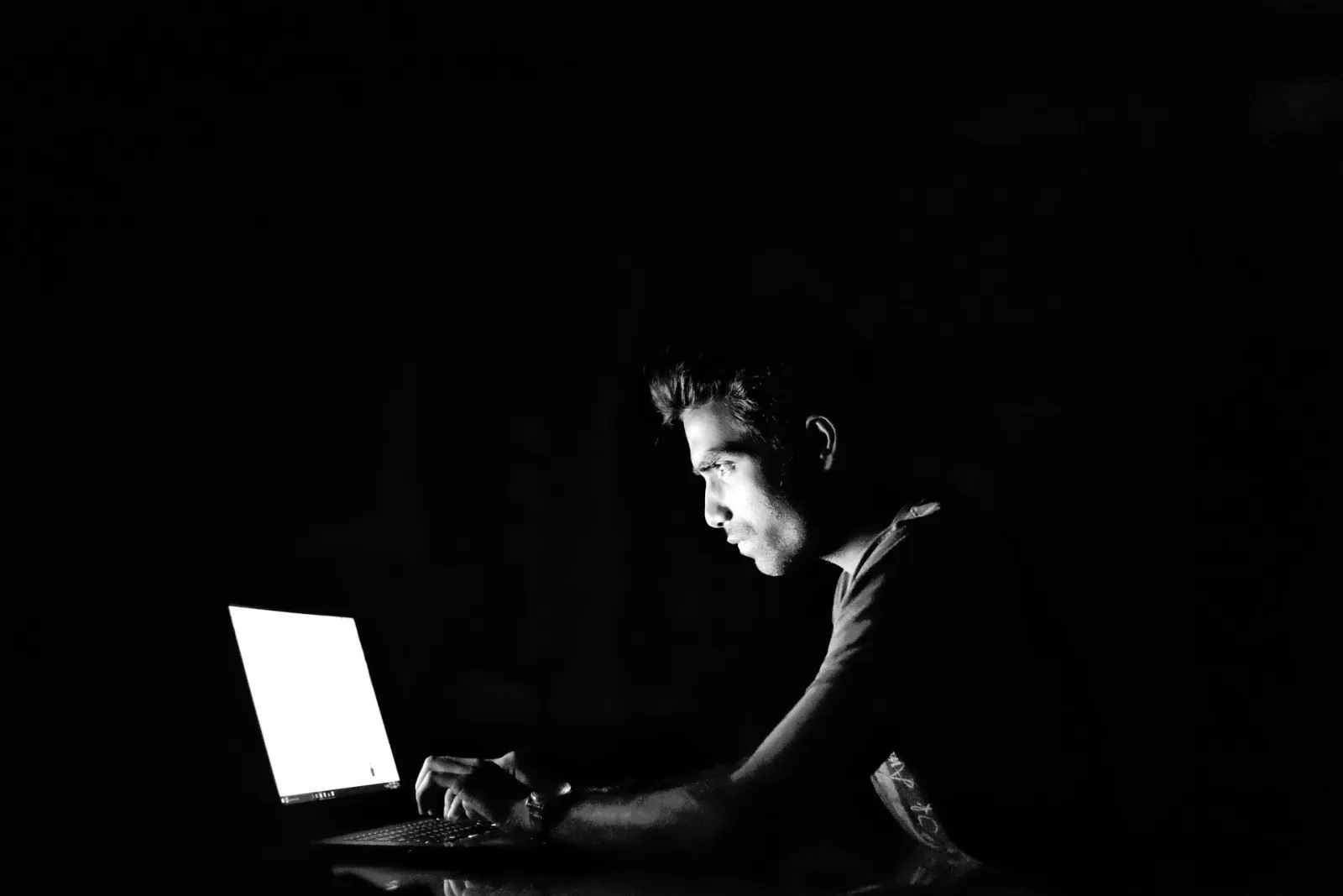 A person working alone on a laptop in a dark room, lit only by the screen